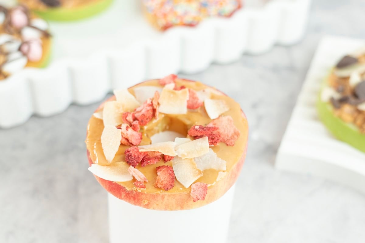 A red apple with core removed cut in a donut shape, spread with peanut butter, topped with freeze dried raspberries and coconut flakes.