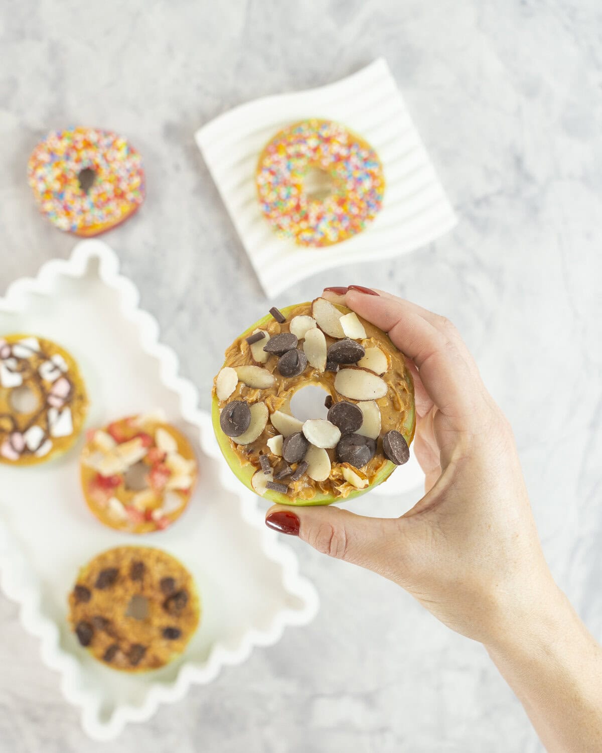 Womans hand holding a green apple with core removed sliced to look like a donut, spread with peanut butter and topped with sliced almonds and chocolate chips. 5 other apple donuts below with various toppings.