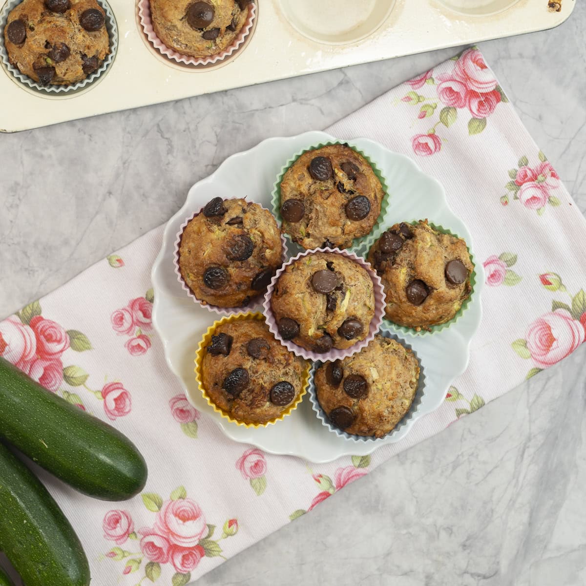 6 muffins stacked on a plate on benchtop on a laid tea towel with pink rose design.