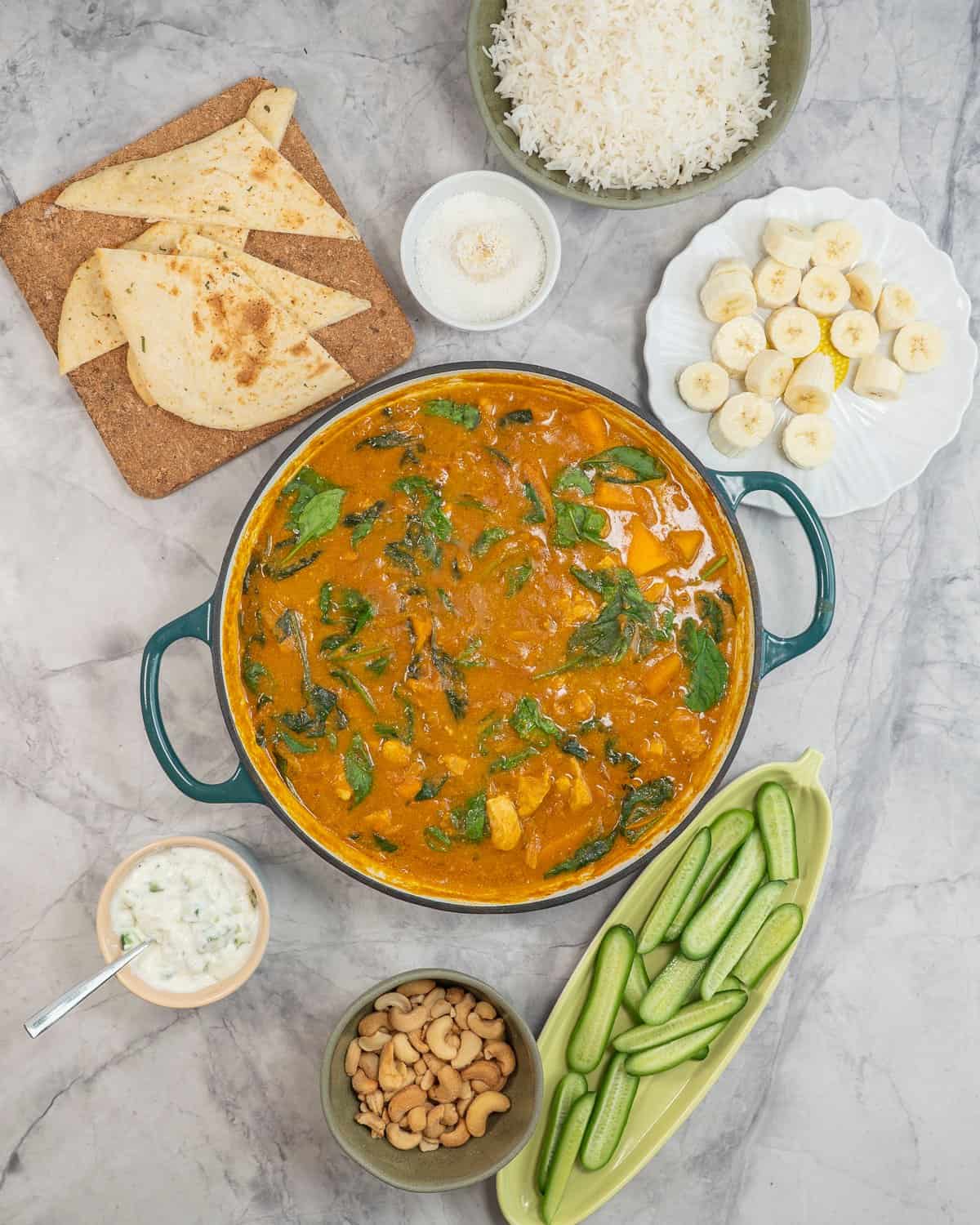 Chicken curry in pan on benchtop with sides, sliced cucumber, cashews, banana slices, naan and dip.