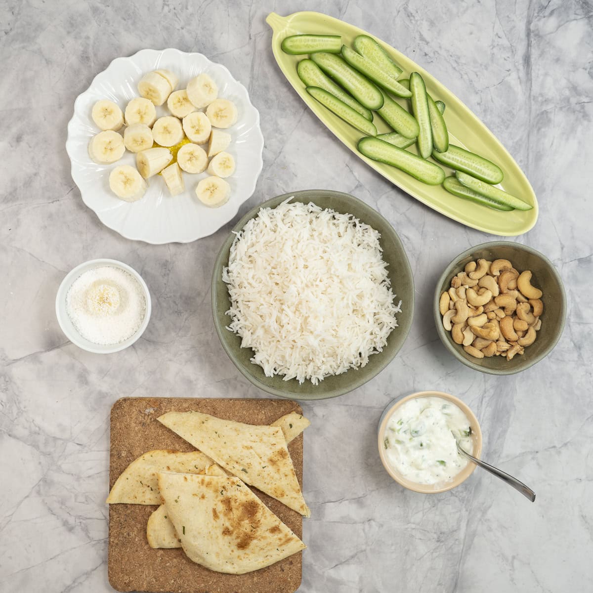 Sliced cucumber, sliced banana, cashews, rice, naan, dip on benchtop to serve with curry.