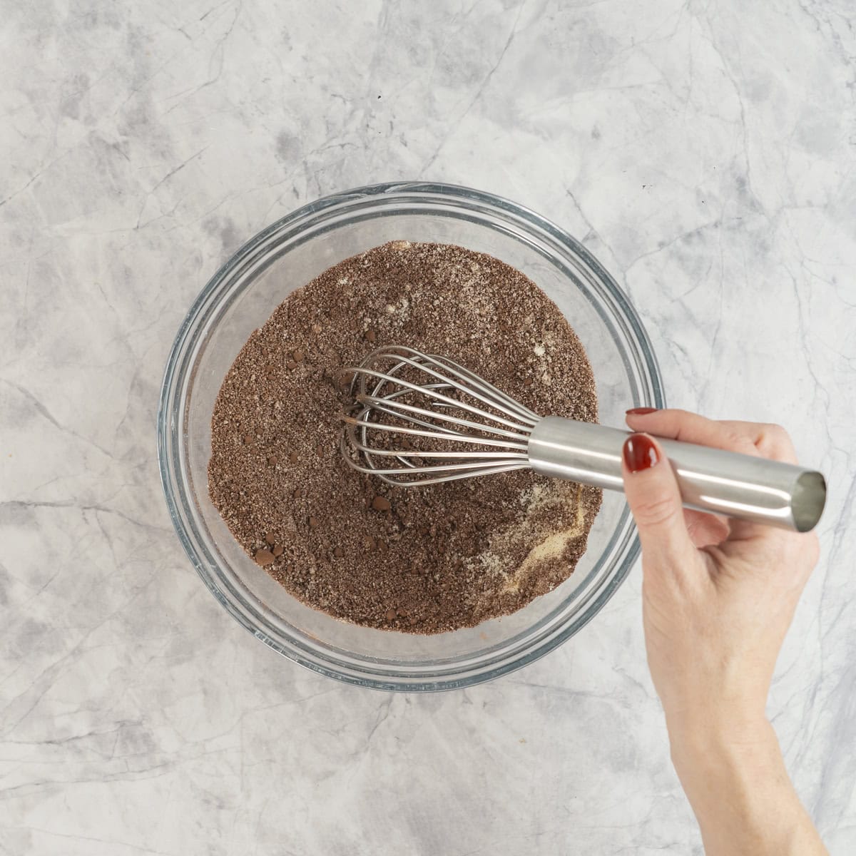 Hand holding a whisk stirring dry ingredients in large glass bowl on benchtop.