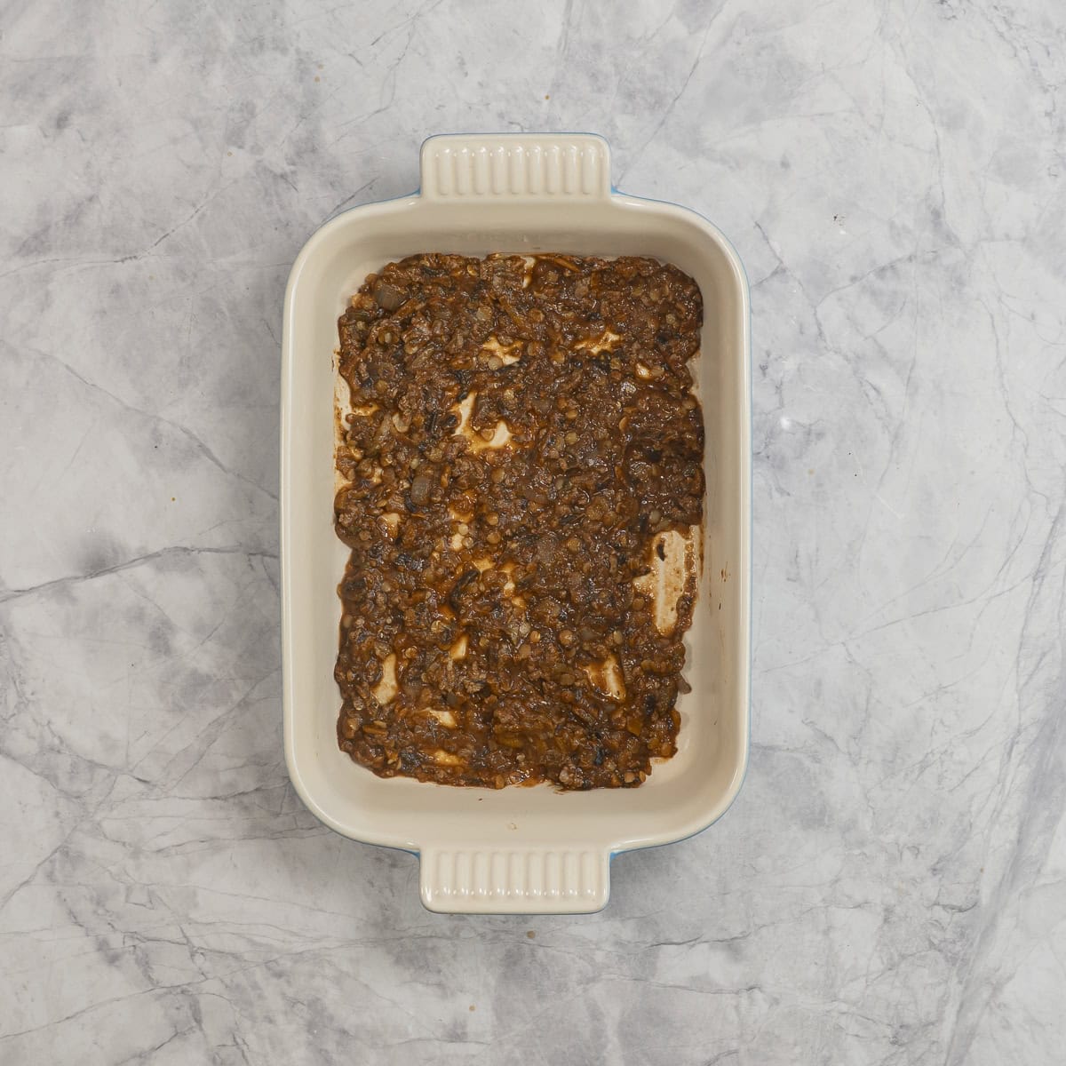 Thin layer of vegetable mince filling on bottom of rectangle baking dish on benchtop.