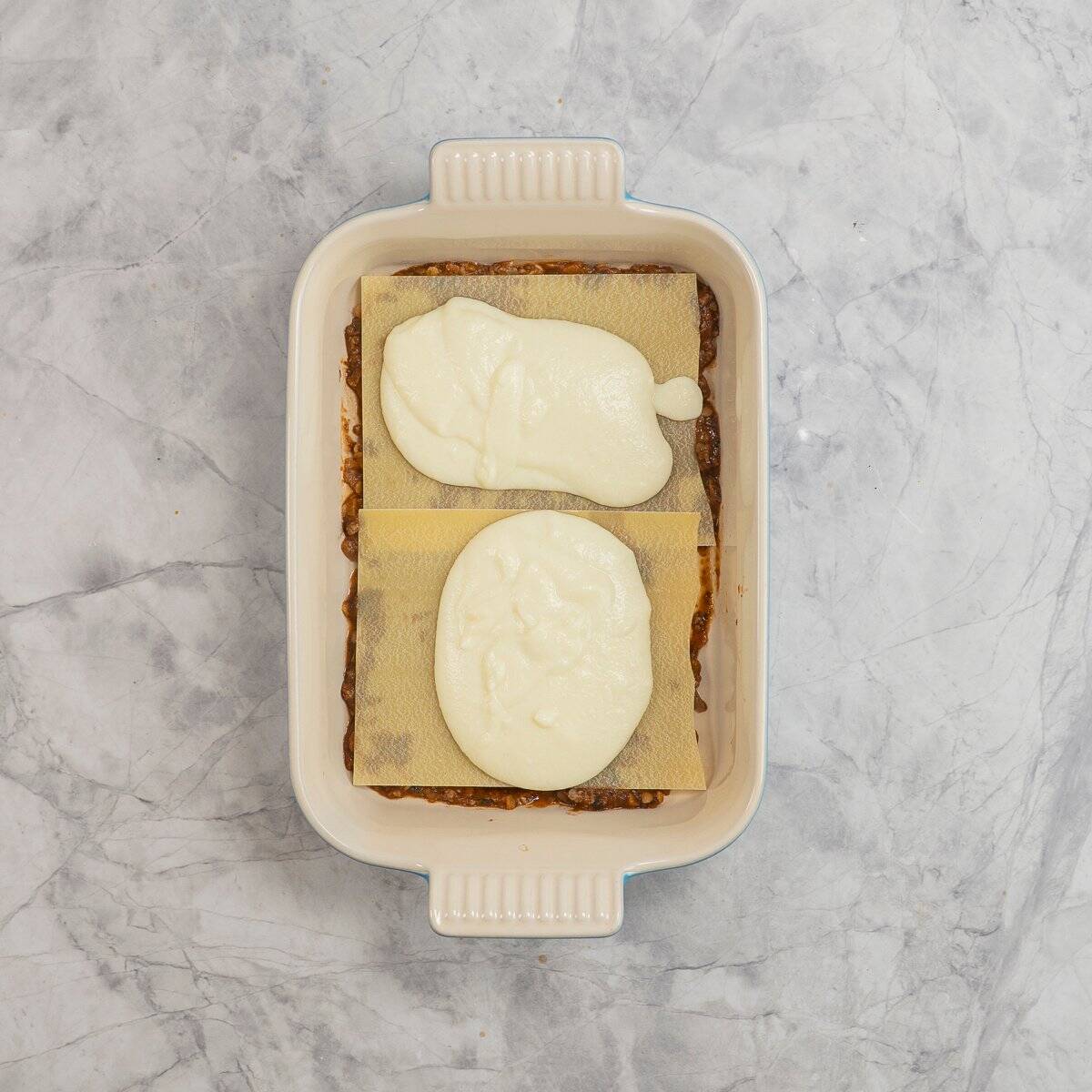 Rectangle baking dish on benchtop with a thin layer of vegetable mince filling, lasagna sheet and two dollops of cauliflower sauce on top.