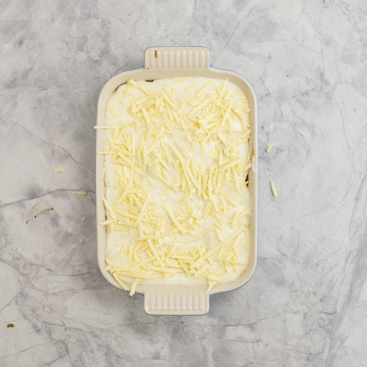 Rectangle baking dish on benchtop with lasagna assembled, cauliflower cheese sauce and sprinkled cheese on top.