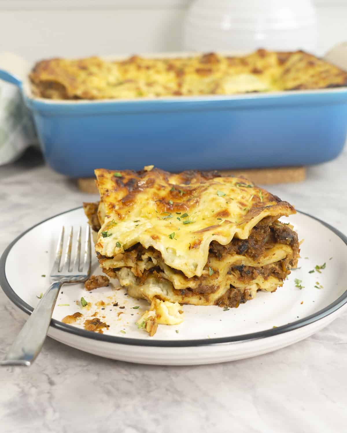 A slice of lasagna showing inside layers of mince, sauce and lasagna sheets on a plate with a fork, Baking dish with lasagna in background.