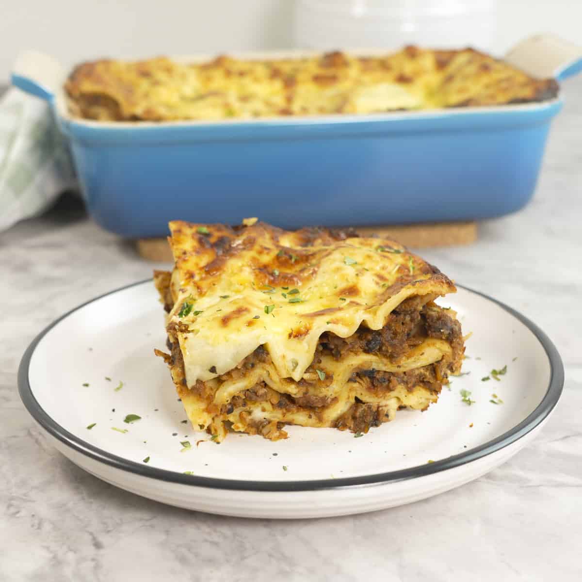 A slice of lasagna showing inside layers of mince, sauce and lasagna sheets on a plate, Baking dish with lasagna in background.