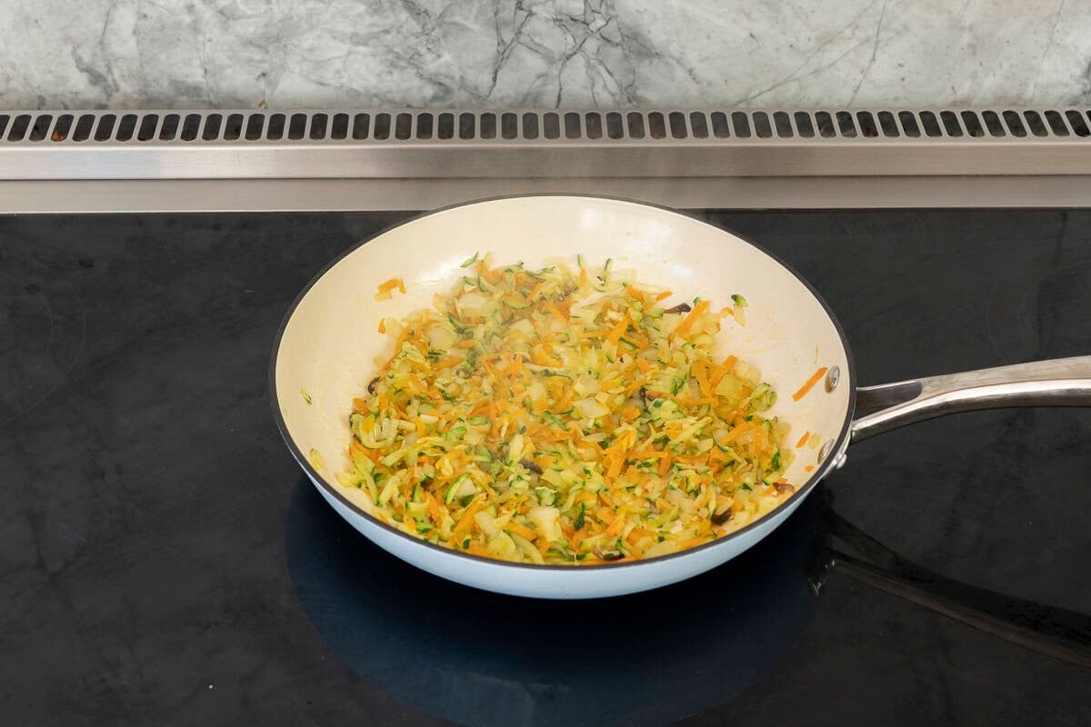 A pan with chopped onion, grated carrot, zucchini and mushrooms on a stove.