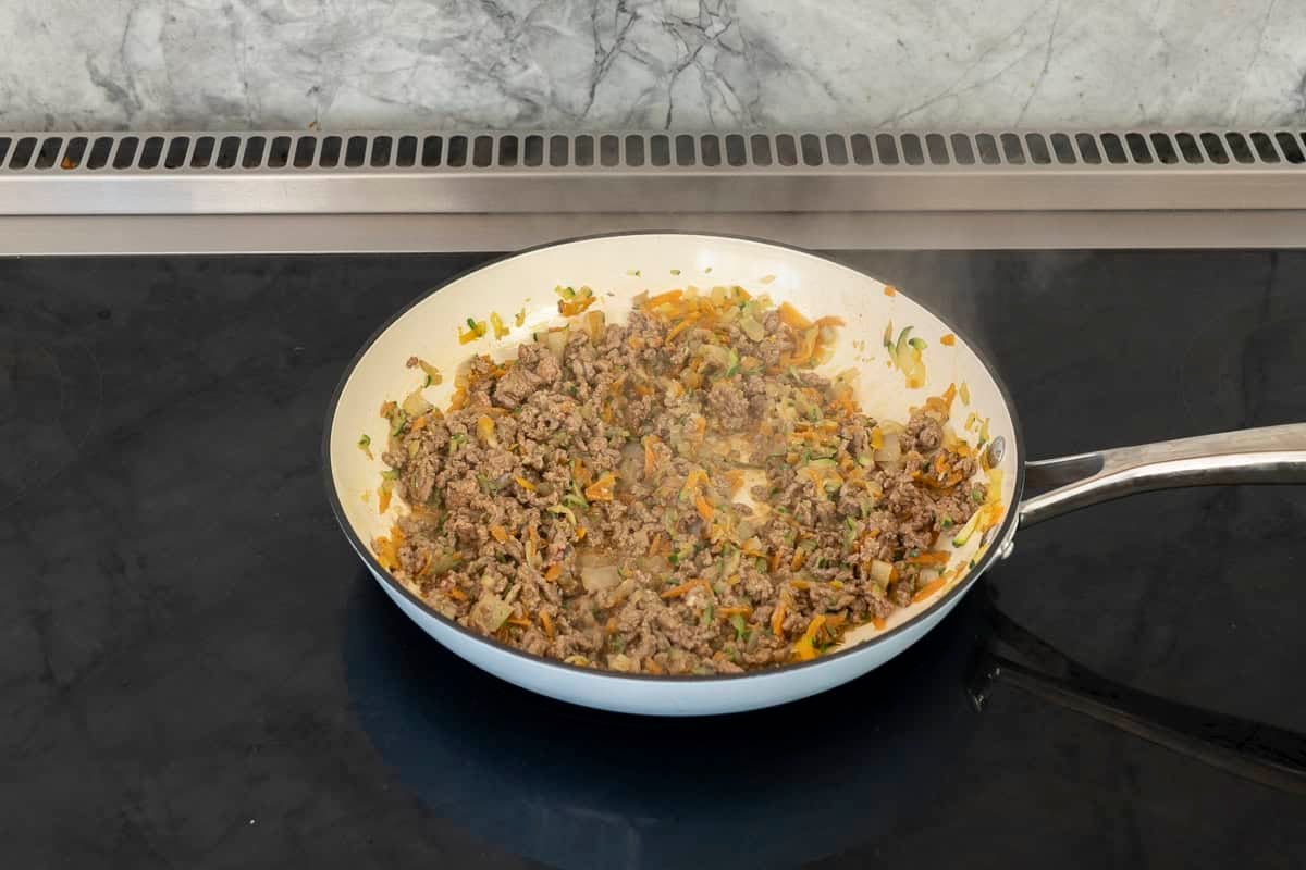 A pan cooking on stove with chopped onion, grated carrot, zucchini and mince.