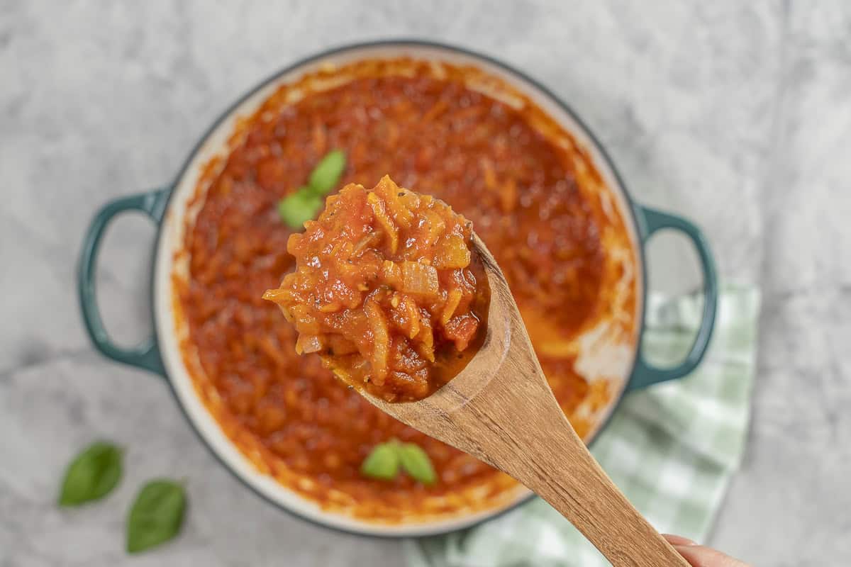 Hand holding a wooden spoon of marinara sauce over a pan of tomato sauce.