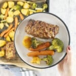 Hand holding a dinner plate with roast carrot, broccoli and potato and a small meatloaf above a lined baking tray of roast vegetables and other mini meatloaf's.