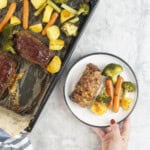 Hand holding a dinner plate with roast carrot, broccoli and potato and a small meatloaf next to a lined baking tray of roast vegetables and other mini meatloaf's.