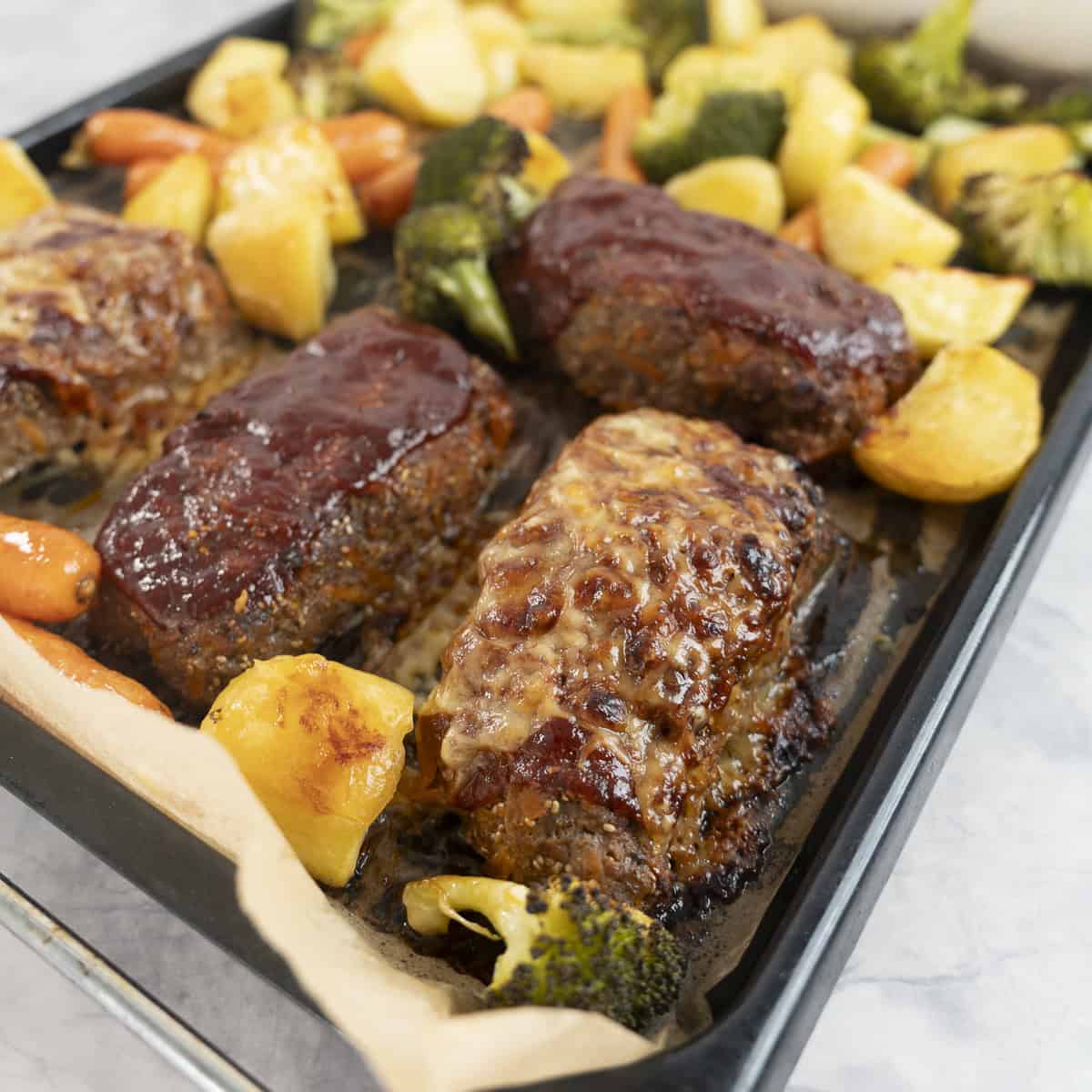Lined baking tray with roasted vegetables and four mini rectangle meatloaf's, with cheese melted on top.