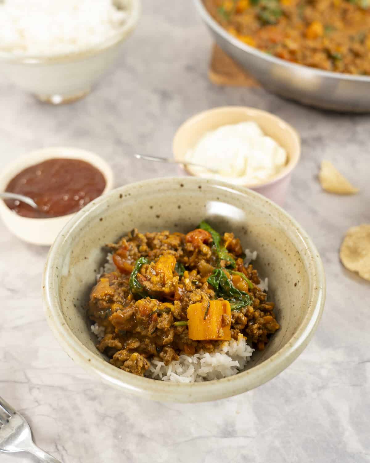A bowl of curry served on rice in a dinner bowl on bench top, small ramekins with