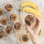 Hand holding a cooked banana chocolate chunk muffin, more below on cooling rack and baking paper on bench top with gooey melted chocolate on top.