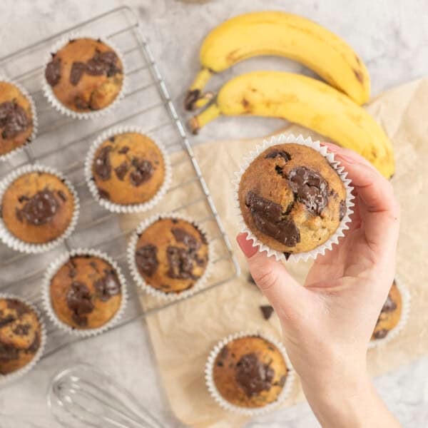 Hand holding a cooked banana chocolate chunk muffin, more below on cooling rack and baking paper on bench top with gooey melted chocolate on top.