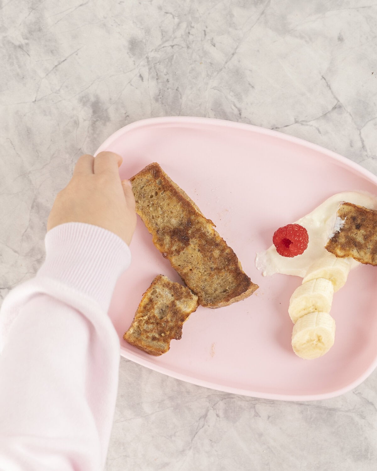 Baby hand reaching for a piece of French toast from a pink baby plate. 