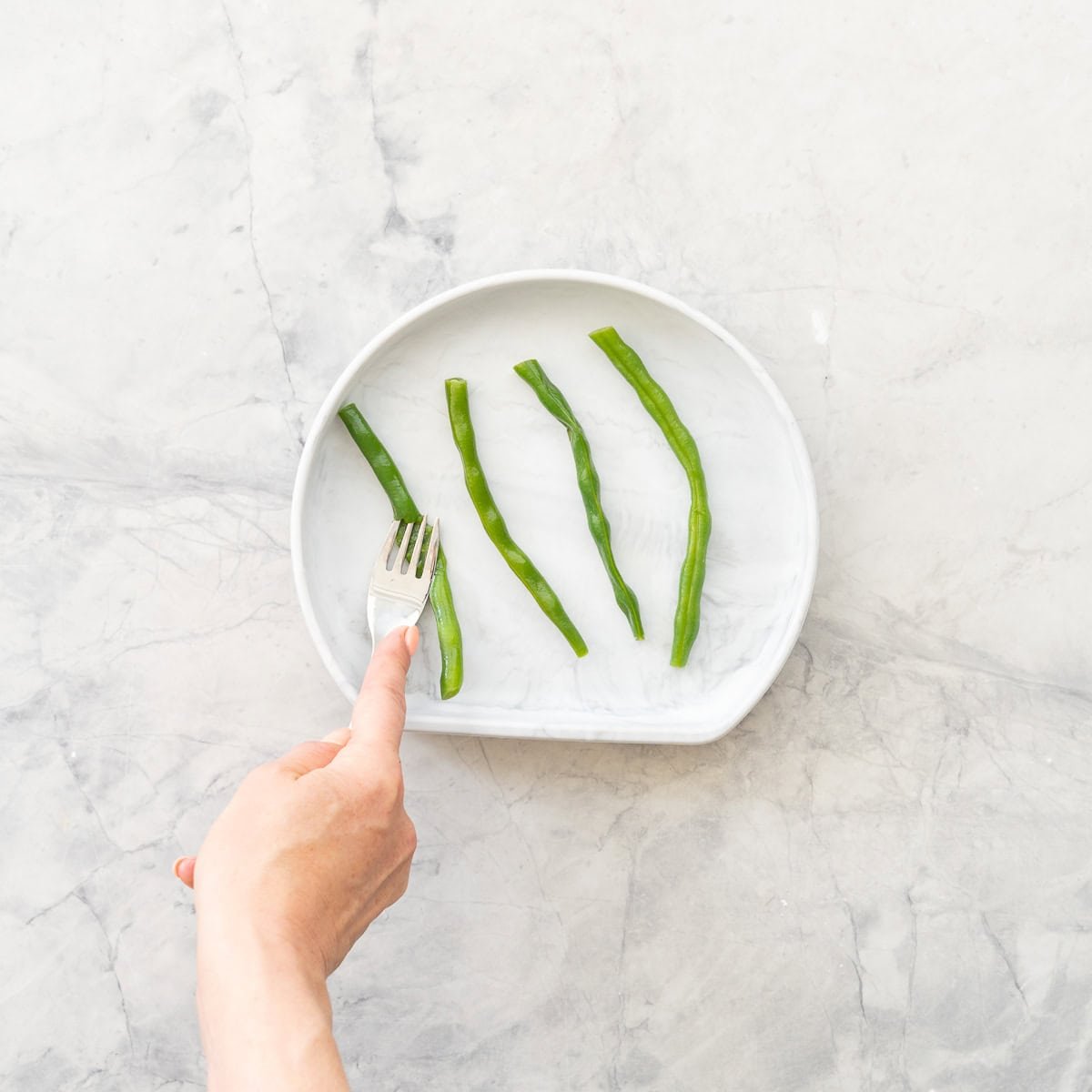 Four cooked green beans on a baby plate, one squashed with a baby fork. 