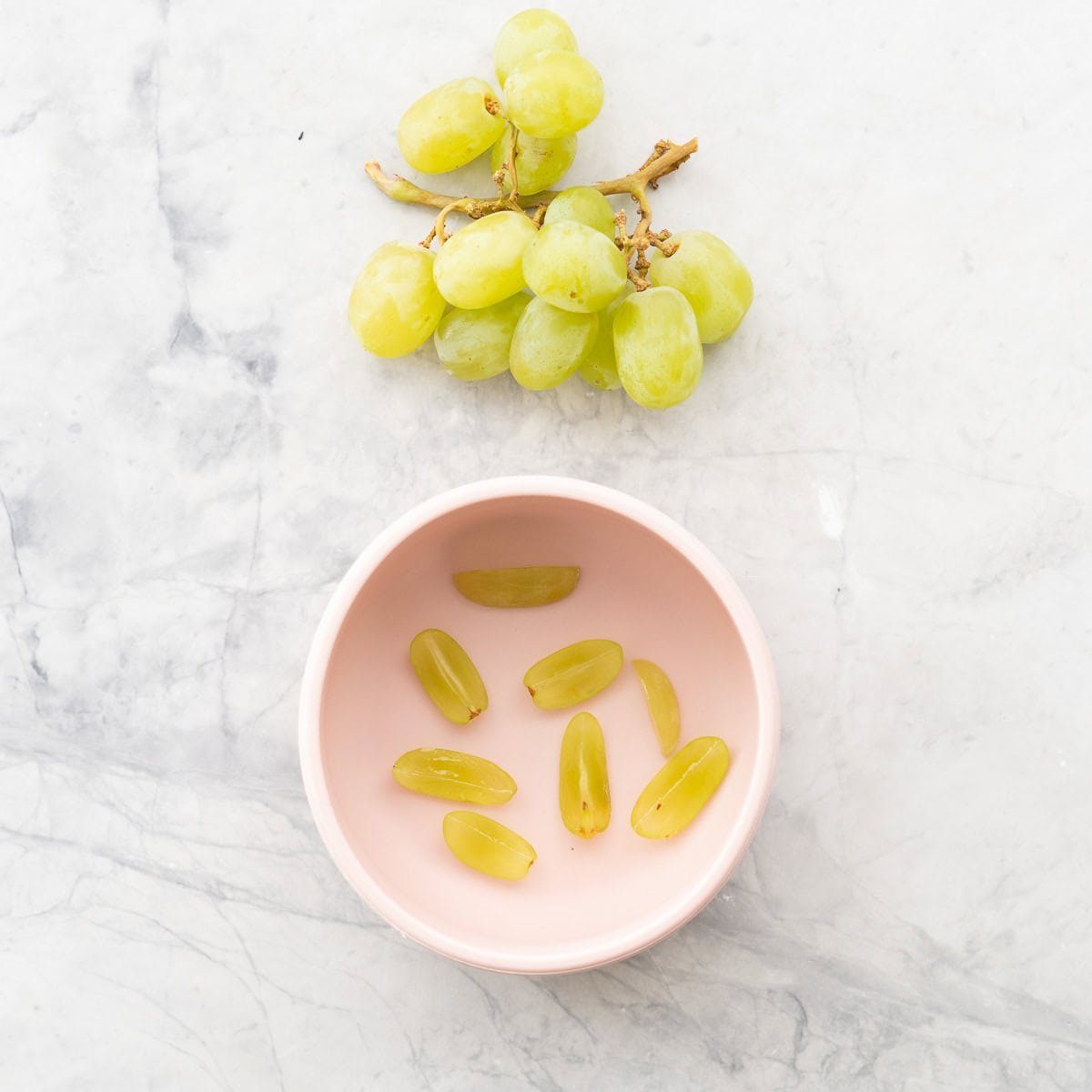 A bunch of green grapes next to a pink baby bowl of grapes cut into quarters lengthways. 