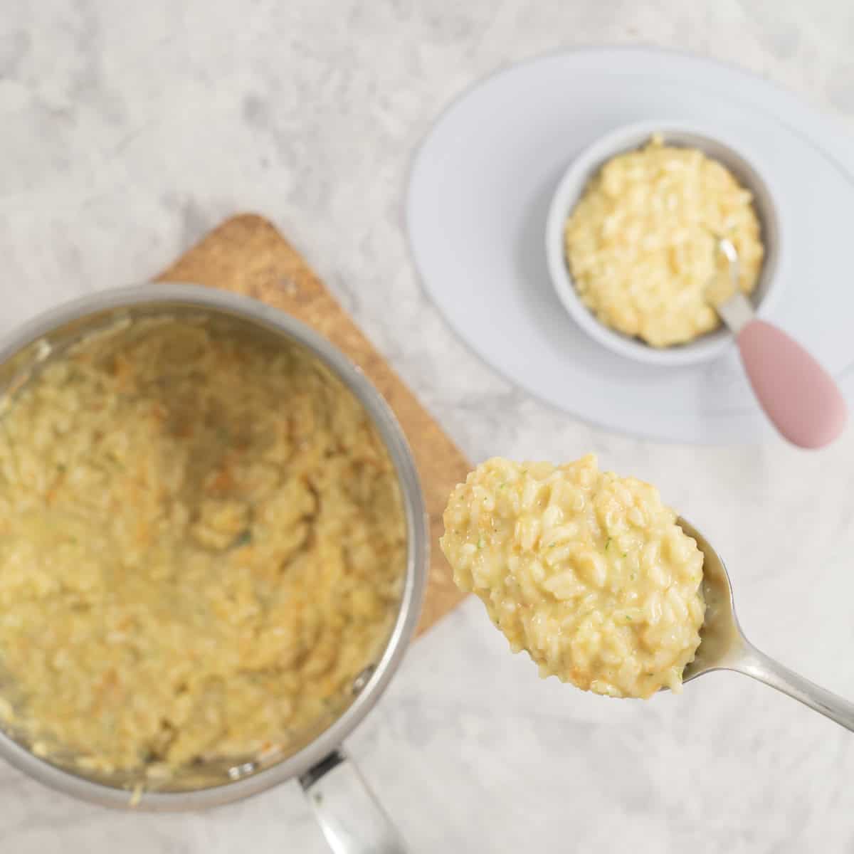 Pot on cork board on bench top with hand holding a serving spoon with scoop of creamy rice on it, next to it a silicon baby bowl with toddler fork inside.