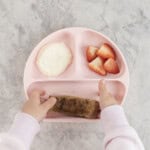 A toddlers hand taking a piece of eggy bread from a pink silicone baby plate with cut strawberries and yogurt.