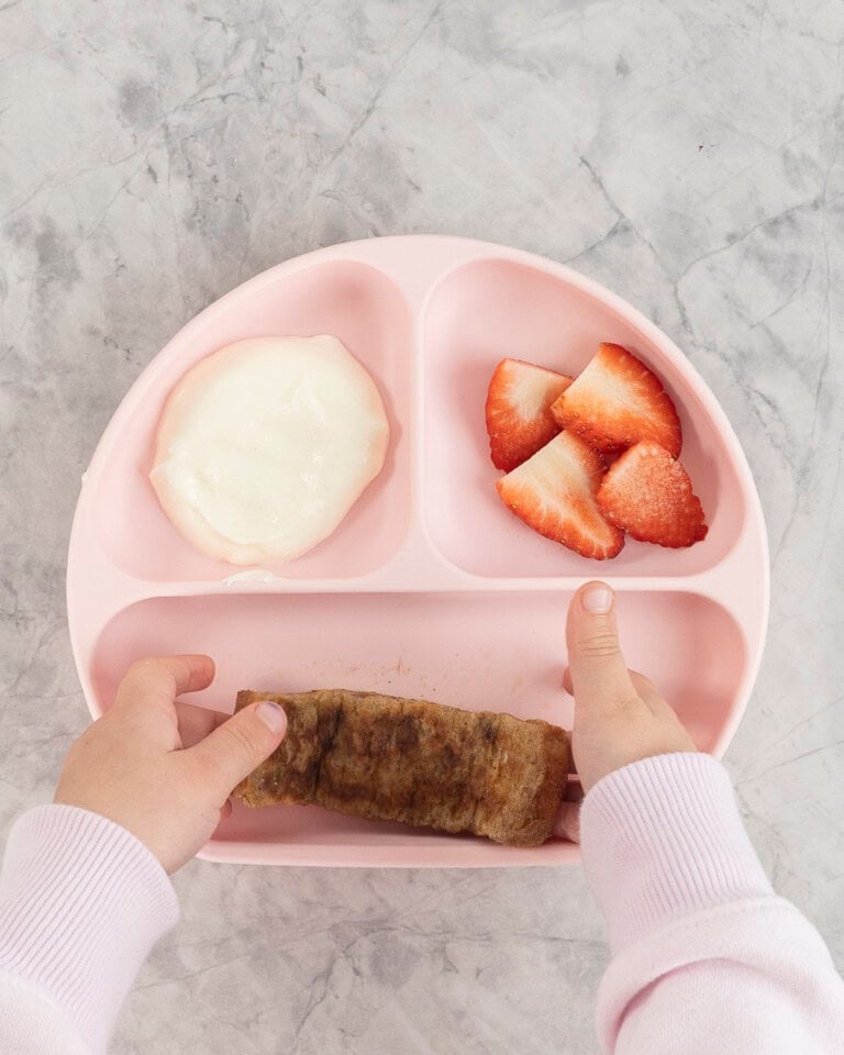 A toddlers hand taking a piece of eggy bread from a pink silicone baby plate with cut strawberries and yogurt.
