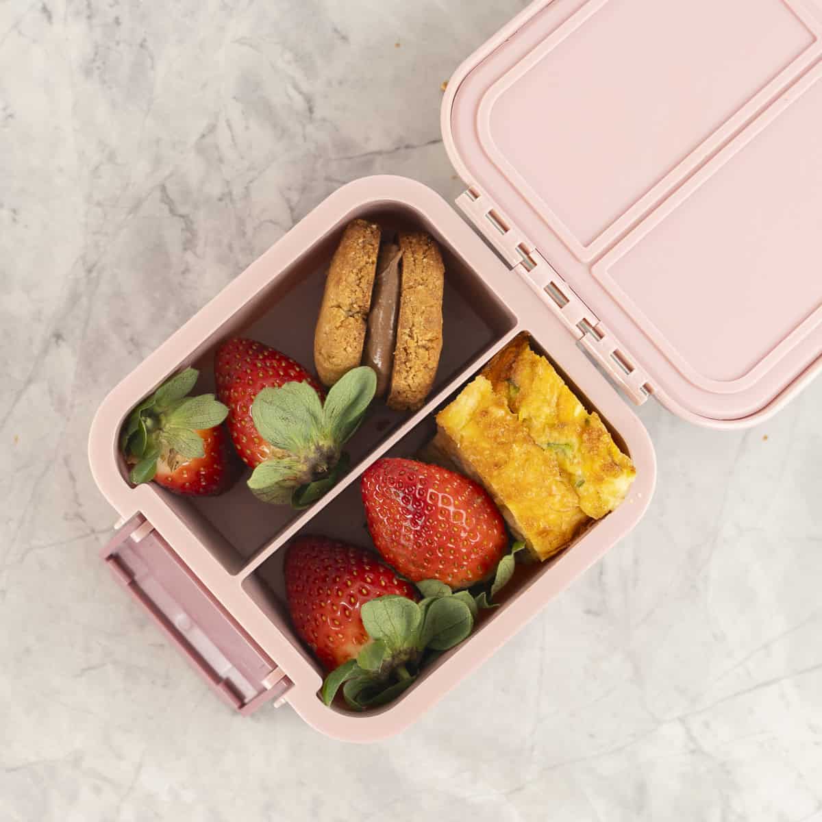 Small pink lunchbox with peanut butter sandwich cookie, strawberries and quiche inside.