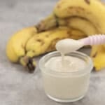 Small glass container with pink baby spoon scooping out banana pudding, bananas stacked in background.