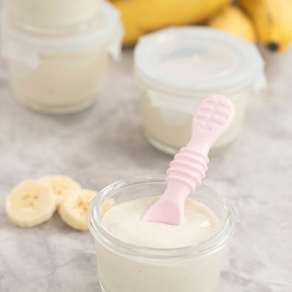 Four small glass containers with banana pudding inside, one with no lid on and a pink baby silicone spoon inside.