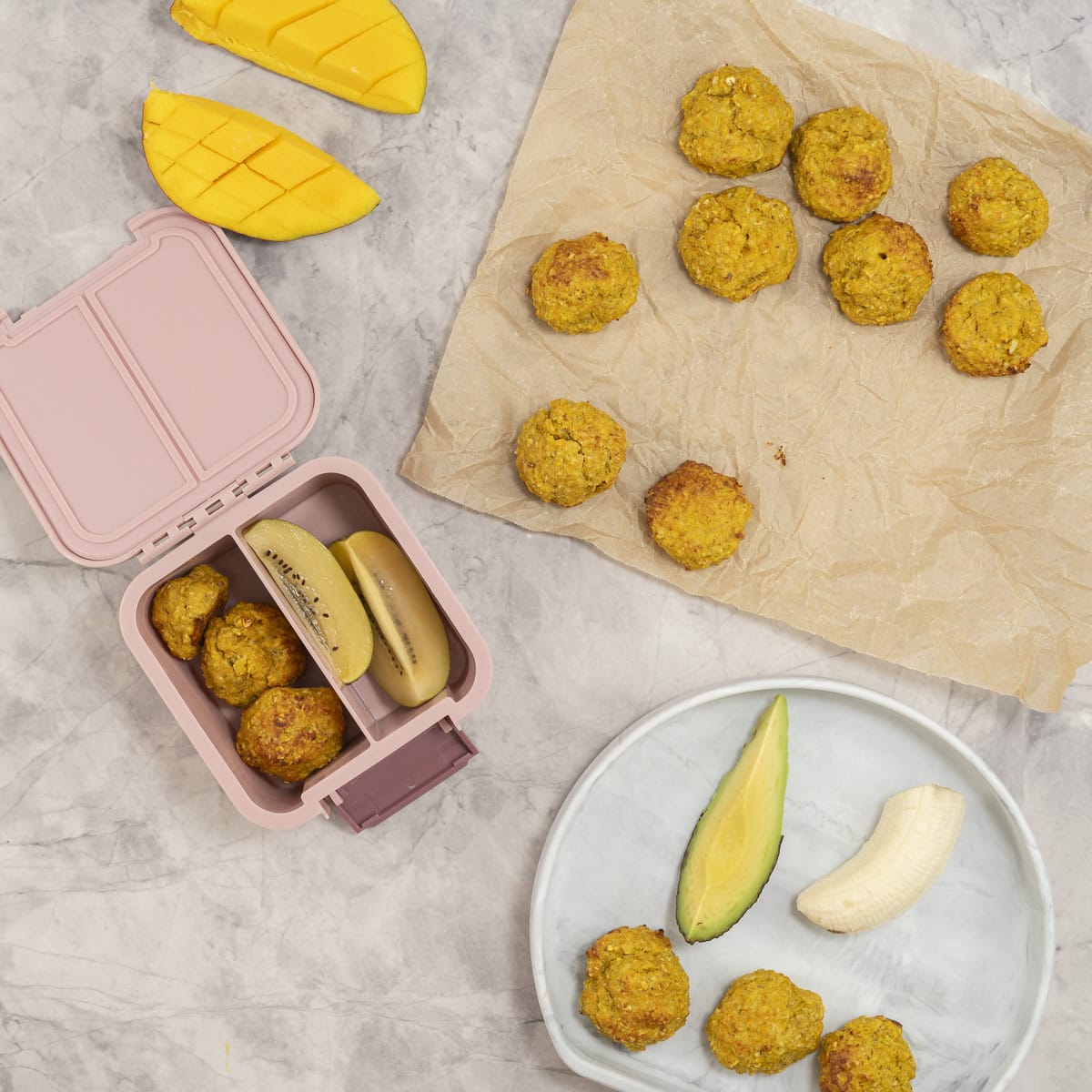 Meatballs on a piece of parchment paper next to a small pink lunchbox packed with meatballs and kiwi.