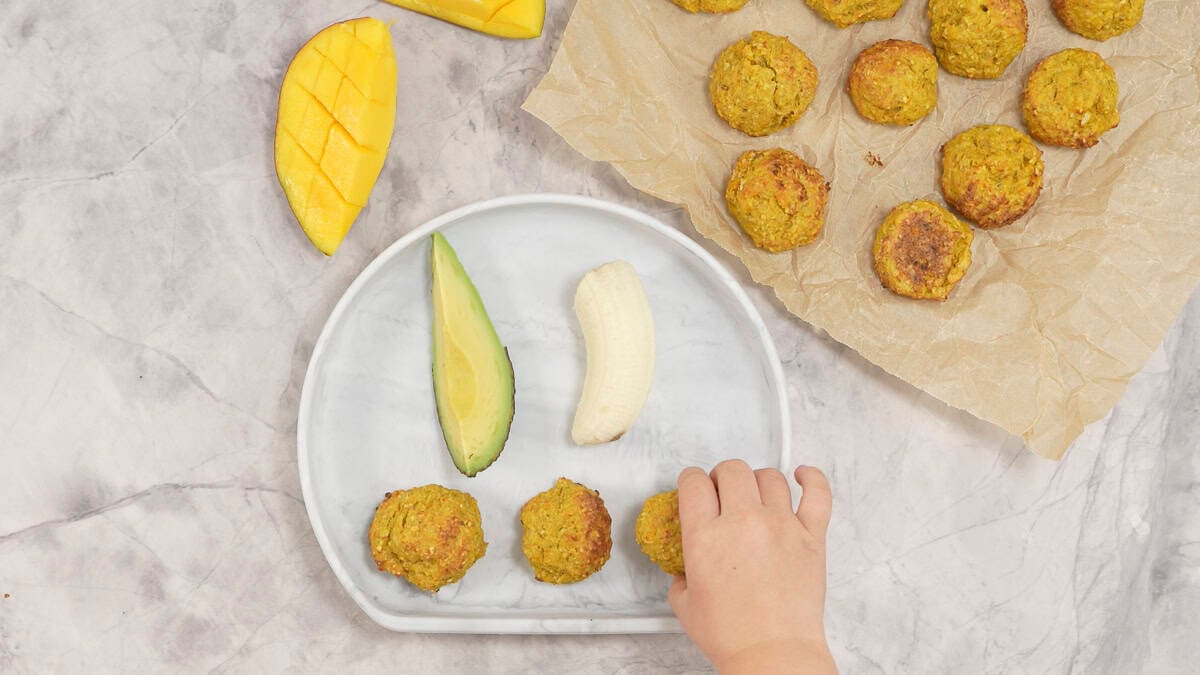 A baby hand reaching for a meatball on a baby plate next to other meatballs and pieces of avocado. 