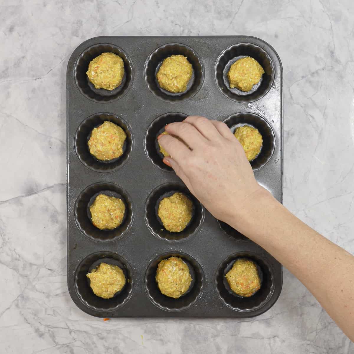 A hand placing a ball of meatball mix into the cavity of a muffin tray, all the other cavities have meatball mix in them. 