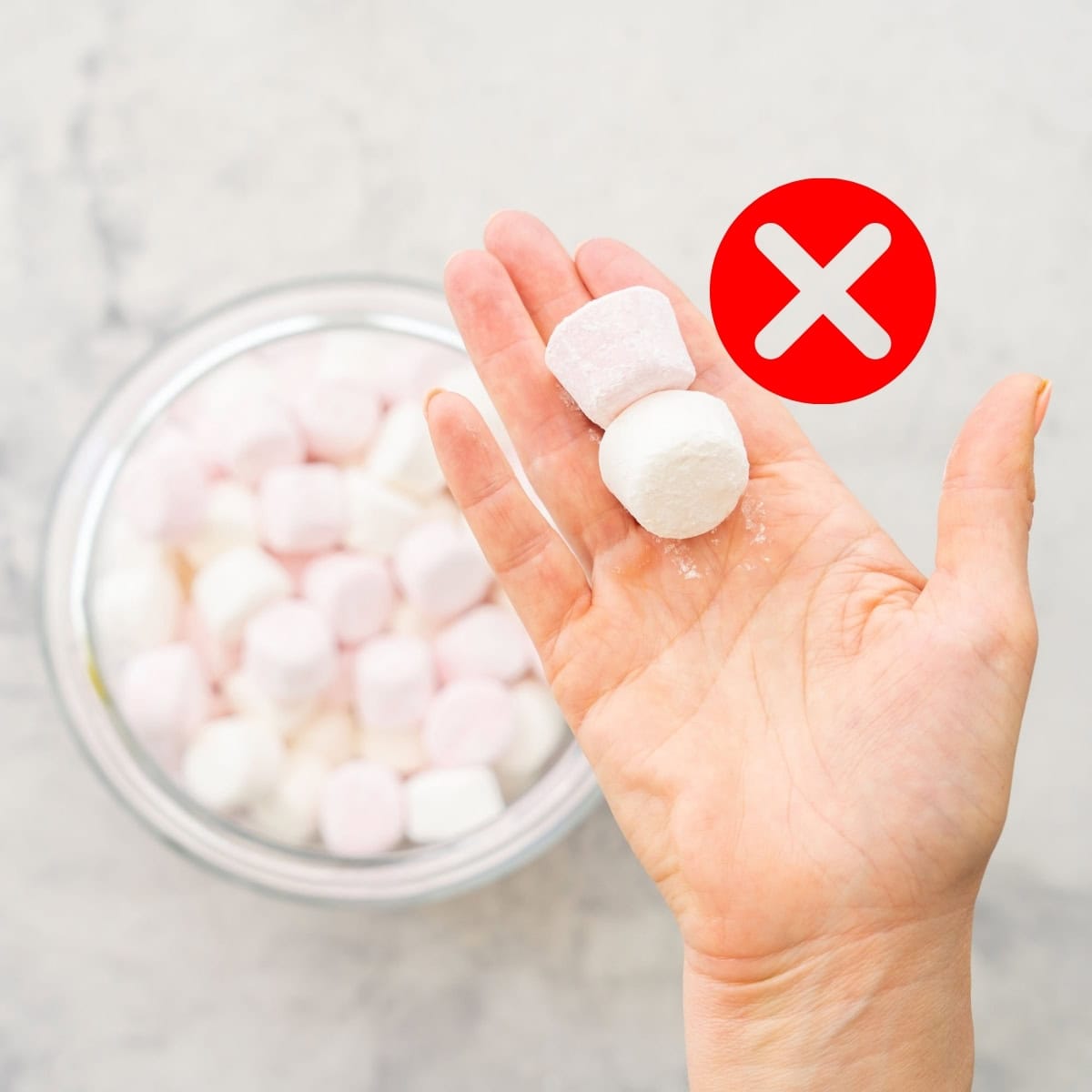 A hand holding 2 marshmallows above a jar of more marshmallows with a Red Cross next it. 