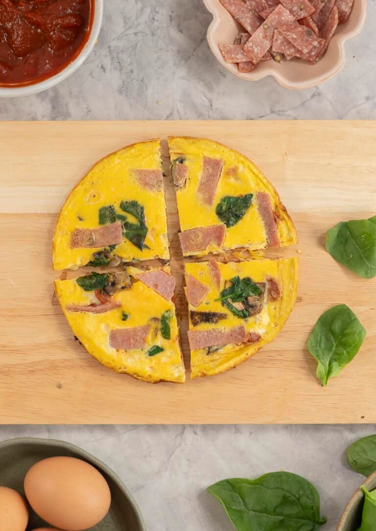 Wooden chopping board with pizza egg cut into four slices, toppings on bench top.