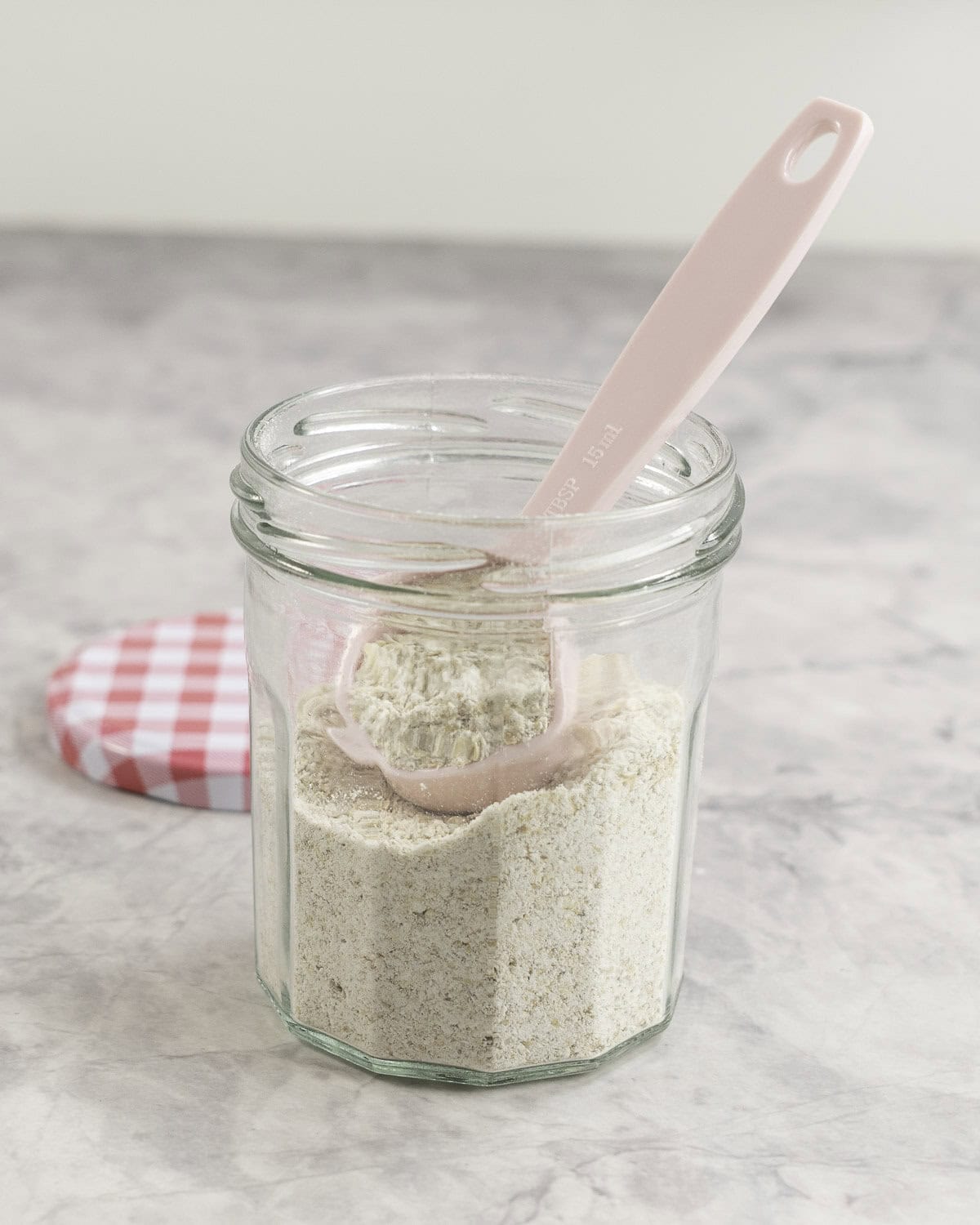 A glass jar of oat flour/baby oatmeal with a pink tablespoon inside. 