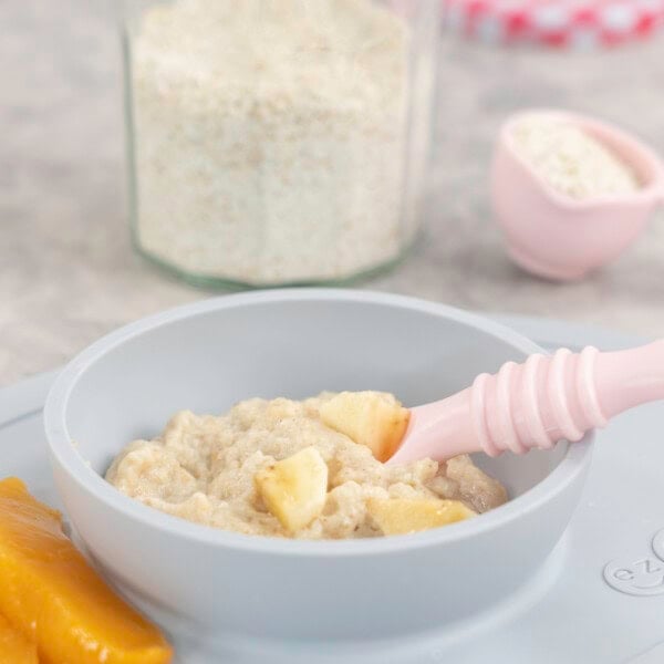 Silicone baby bowl and placemat on marble bench top with baby oats inside, topped with chopped banana pieces with baby spoon inside and two slices of canned peaches on the side, jar of ground oats on benchtop.