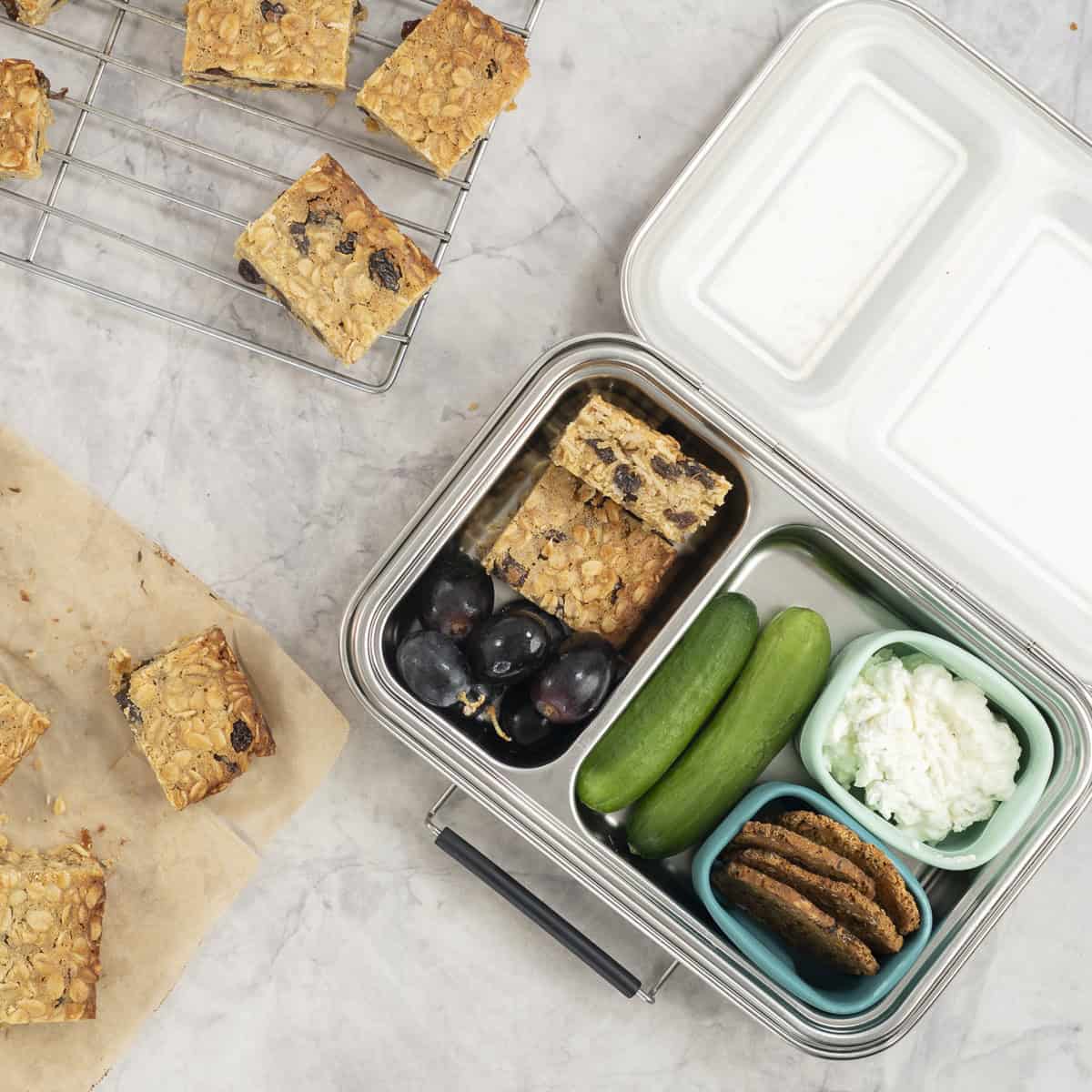 Stainless steel lunchbox on benchtop with oat slice, red grapes, mini cucumbers, crackers and cottage cheese in compartments.