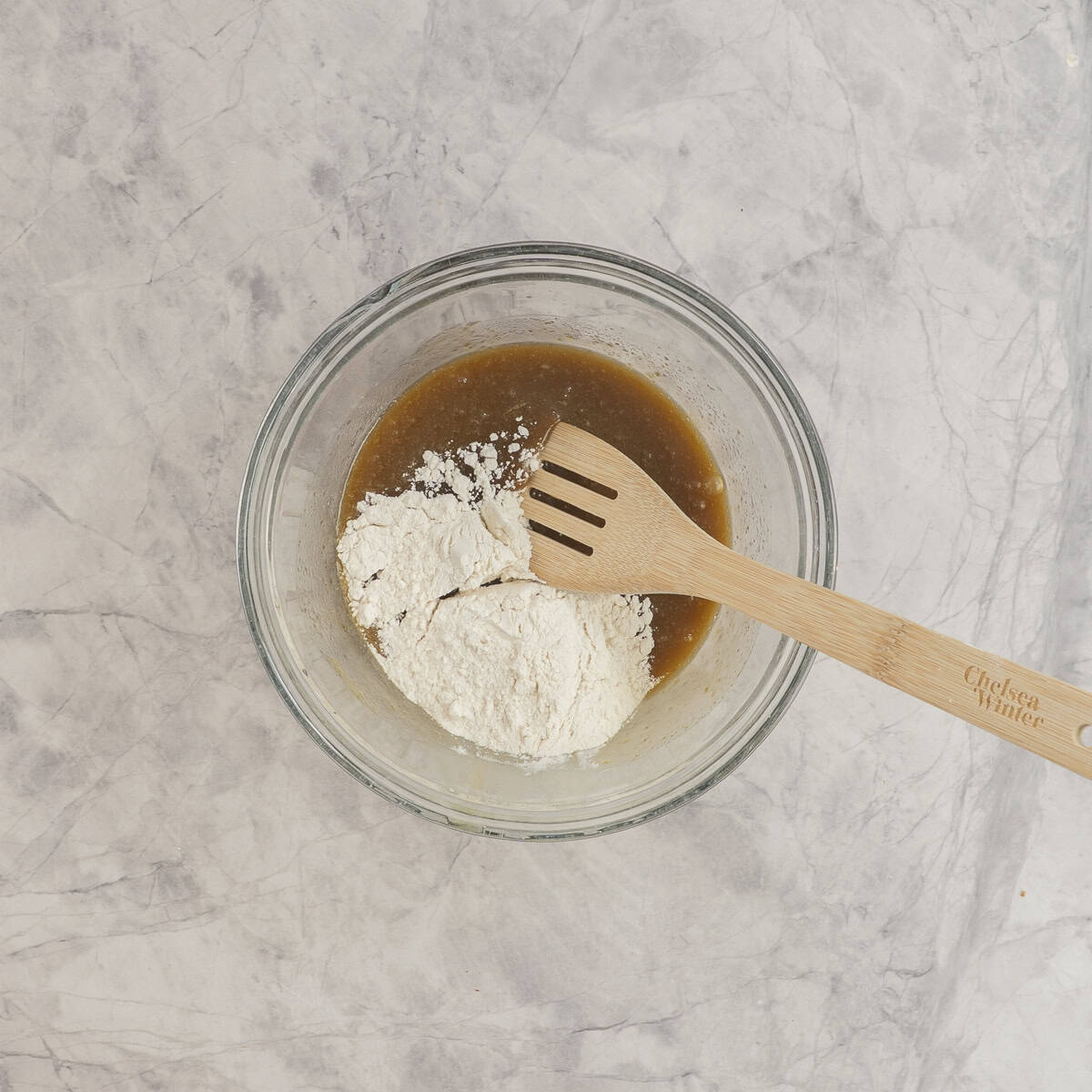 Flour added into wet ingredients in glass bowl on benchtop with wooden spoon inside.