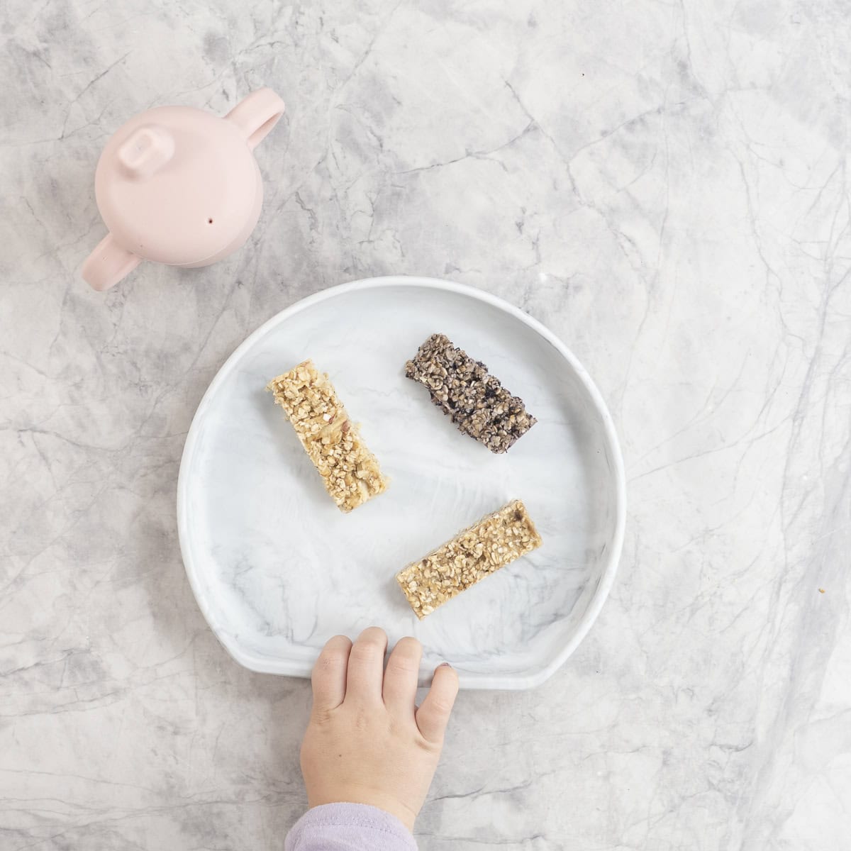 Toddler hand reaching for a marble silicone plate with three porridge fingers and pink silicone cup on benchtop.