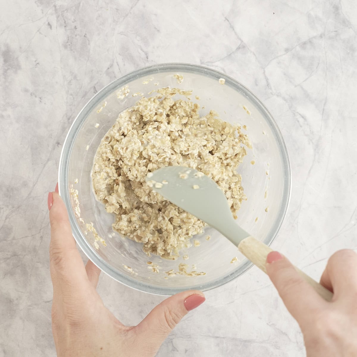 Womans hand holding a spatula mixing together milk and oats in a glass bowl.