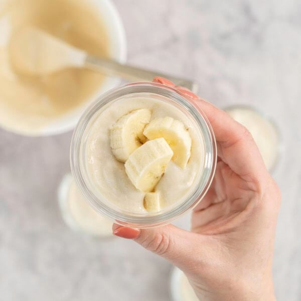 Woman's hand holding up a small glass ramekin with yogurt and banana slices inside, more filled ramekins below on bench top and white bowl with spatula inside.