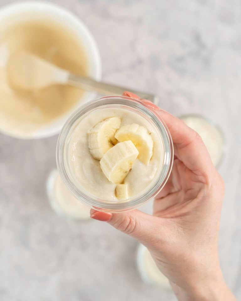 Woman's hand holding up a small glass ramekin with yogurt and banana slices inside, more filled ramekins below on bench top and white bowl with spatula inside.