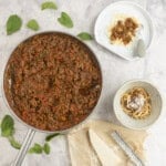 Pan on marble benchtop with bolognese mince inside, a dinner plate and bowl served with spaghetti pasta, spinach leaves scattered on bench top.
