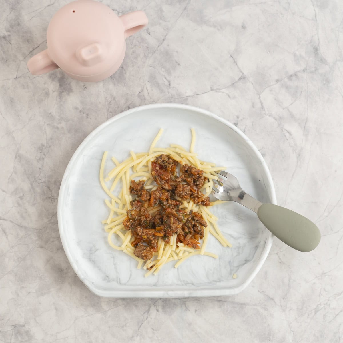 Silicone baby plate on marble bench top with bolognese on spaghetti, baby spoon and cup next to plate.