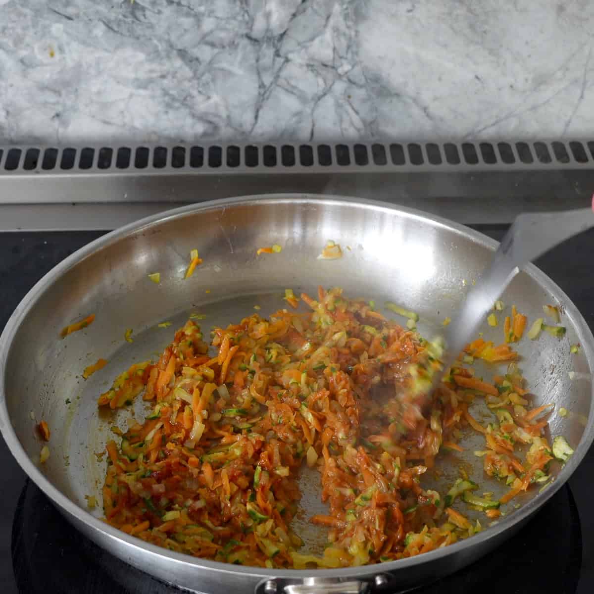 Grated carrot, zucchini, and onion in a large pan on stove top being mixed with metal spatula.
