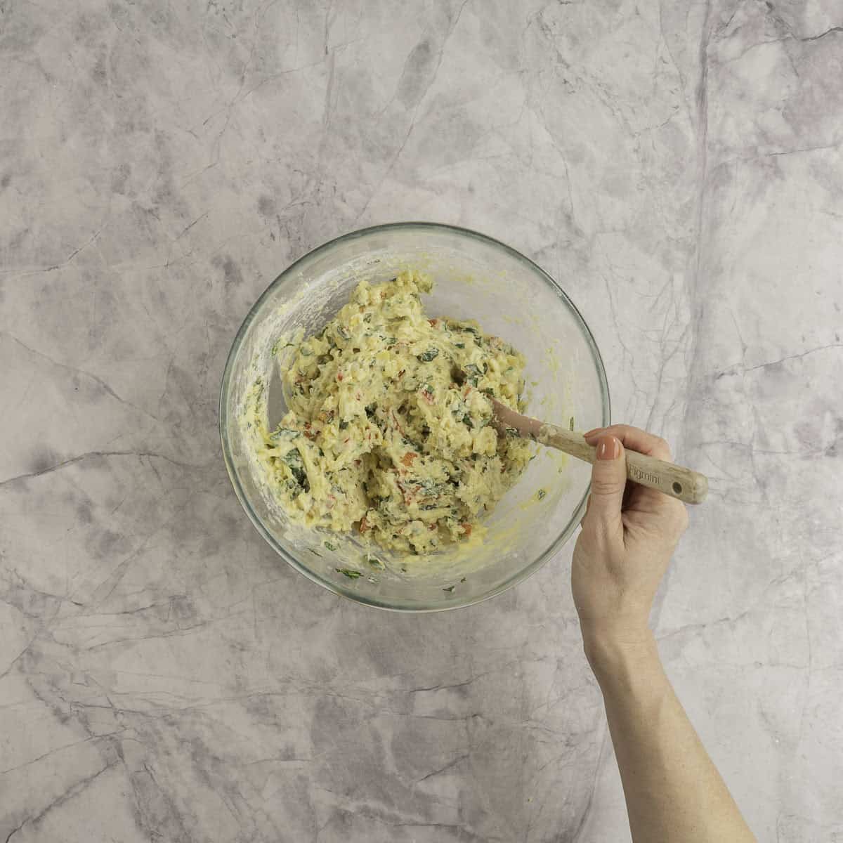 Vegetable mixture mixed together with a spatula inside a large glass bowl on bench top.