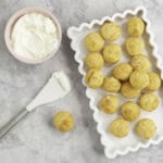 White rectangle dish with wavy border, batch of mini muffins inside, small pink ramekin with greek yogurt inside and a spatula on bench top.