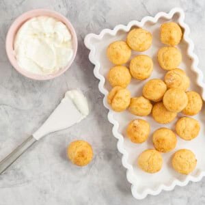 White rectangle dish with wavy border, batch of mini muffins inside, small pink ramekin with greek yogurt inside and a spatula on bench top.