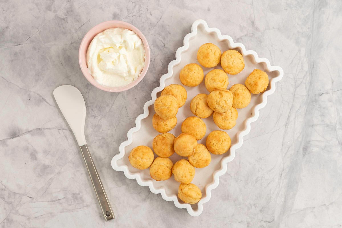 White rectangle dish with wavy border, batch of mini muffins inside, small pink ramekin with greek yogurt inside and a spatula on bench top.
