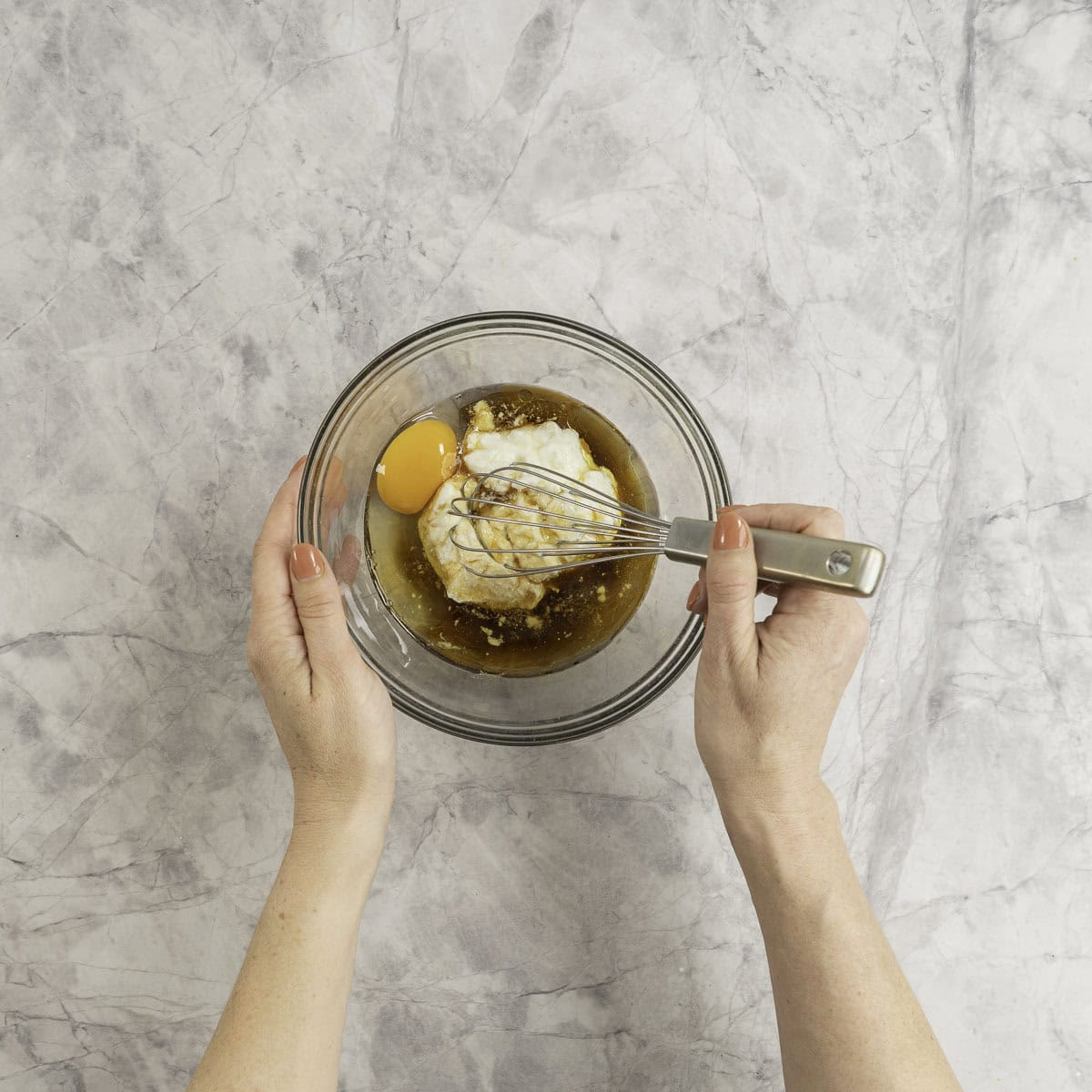 A small glass bowl on marble bench top with wet ingredients inside, woman's hand holding a whisk stirring.